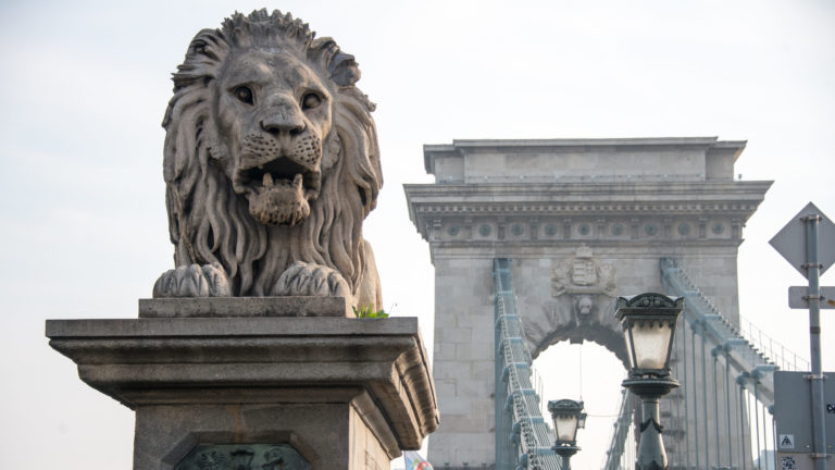 Stone lion at the Chain Bridge with the pale bridge portal and suspension structure aligned behind it.
