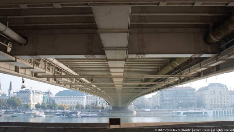 View beneath Elisabeth Bridge showing steel beams overhead, the Danube, riverboats, and Pest buildings beyond.