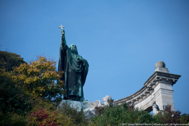 Bronze Saint Gerard figure raising a cross against blue sky, with trees and part of the stone colonnade below.