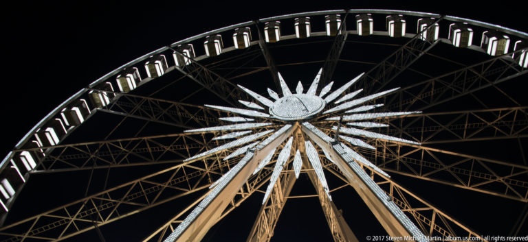 Illuminated hub, spokes, and passenger cabins of the Budapest Eye Ferris wheel against a black night sky.