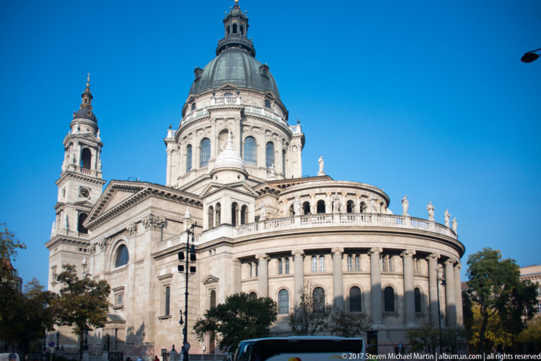 Three-quarter exterior view of St. Stephen’s Basilica with central dome, bell tower, columns, and curved façade.