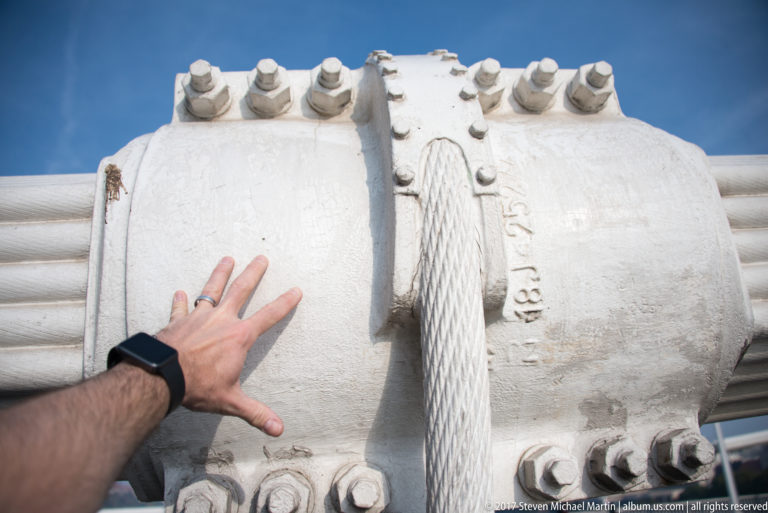 Hand touching a large white-painted bridge cable clamp with heavy bolts and bundled suspension cables.