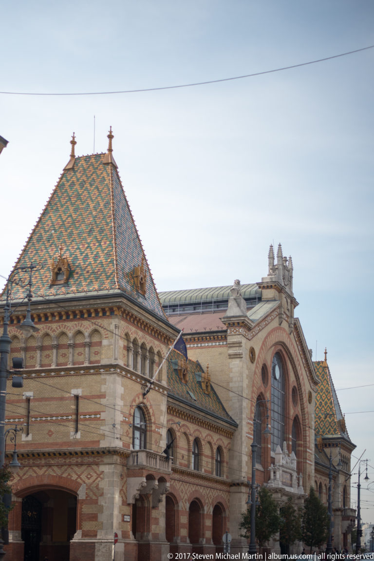Exterior of Budapest's Great Market Hall with brick-and-stone façade, large arched windows, and colorful tiled roof.