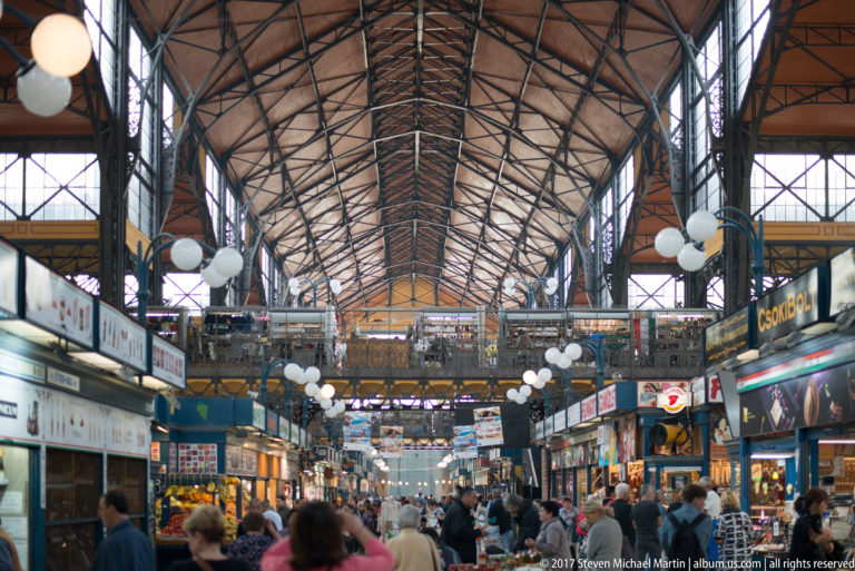 Crowded aisle inside Budapest's Great Market Hall with food stalls, globe lamps, iron roof trusses, and upper gallery.