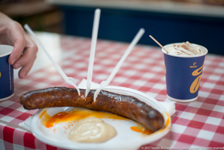 Grilled sausage on a white plate with mustard and plastic forks on a red-and-white checked tablecloth.