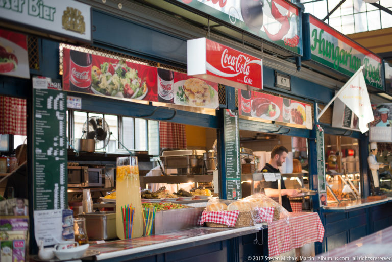 Prepared-food counter in Budapest's Great Market Hall with menu boards, drink dispensers, trays, and checked coverings.