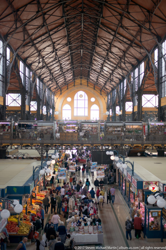 Elevated interior view of Budapest's Great Market Hall with iron roof trusses, upper galleries, and crowded stalls below.