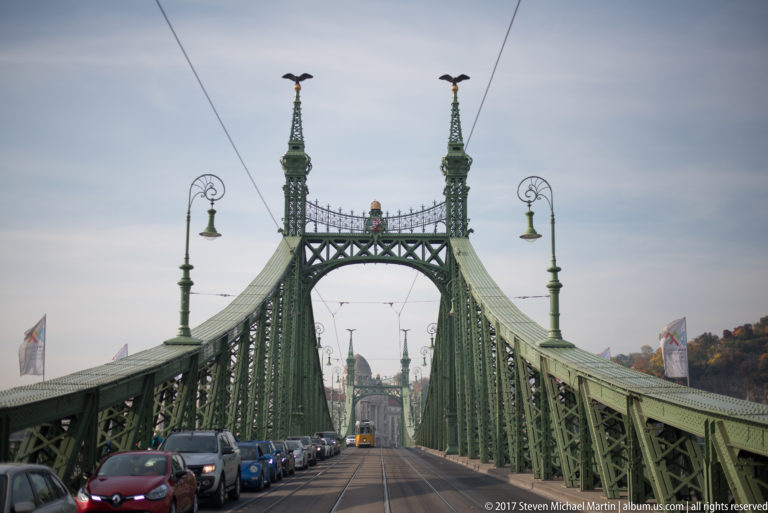 View along Liberty Bridge with cars, tram tracks, overhead wires, ornate lamps, and green steel towers.