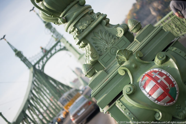 Budapest coat of arms mounted on ornate green ironwork of Liberty Bridge, with the bridge structure blurred behind.