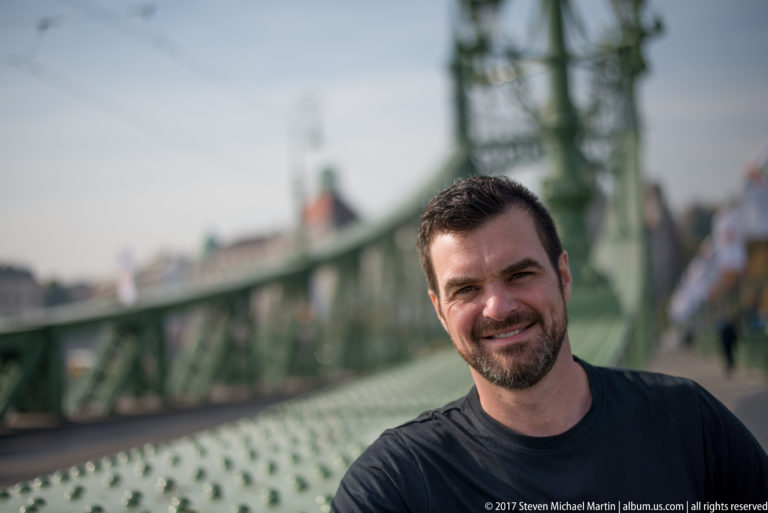 Dark-haired man smiling in front of the green steel structure of Liberty Bridge in Budapest.