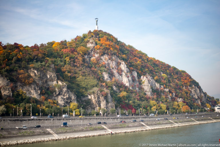 Rocky, tree-covered Gellért Hill in autumn with Budapest's Liberty Statue visible at the summit.