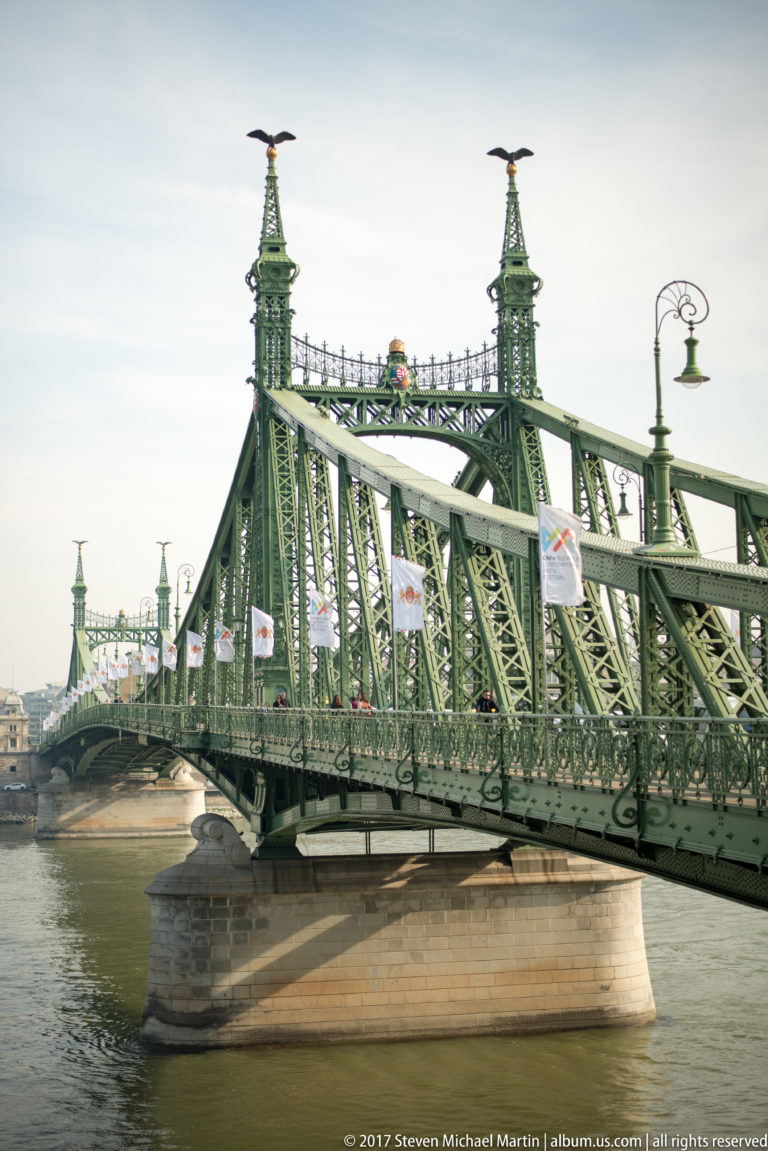 Close side view of Budapest's green Liberty Bridge, showing steel trusses, ornate towers, lamps, and a stone pier.