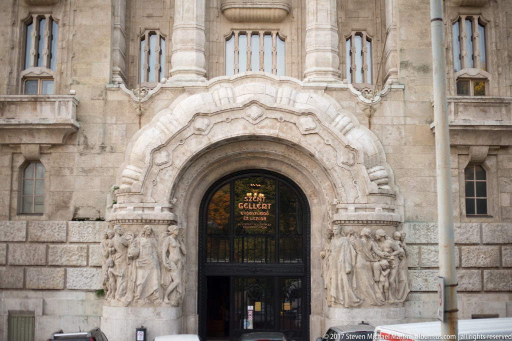 Ornate arched entrance to Gellért Baths with sculptural reliefs, stone columns, and gold lettering above the doorway.
