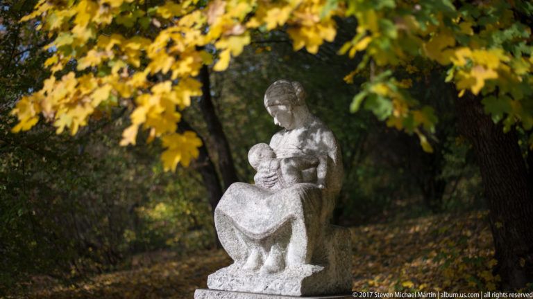 Pale stone sculpture of a seated woman holding a child beneath yellow autumn leaves in a wooded setting.