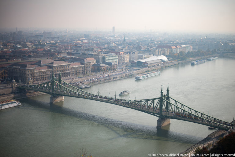 Elevated view of green Liberty Bridge crossing the Danube toward the hazy Pest riverfront.