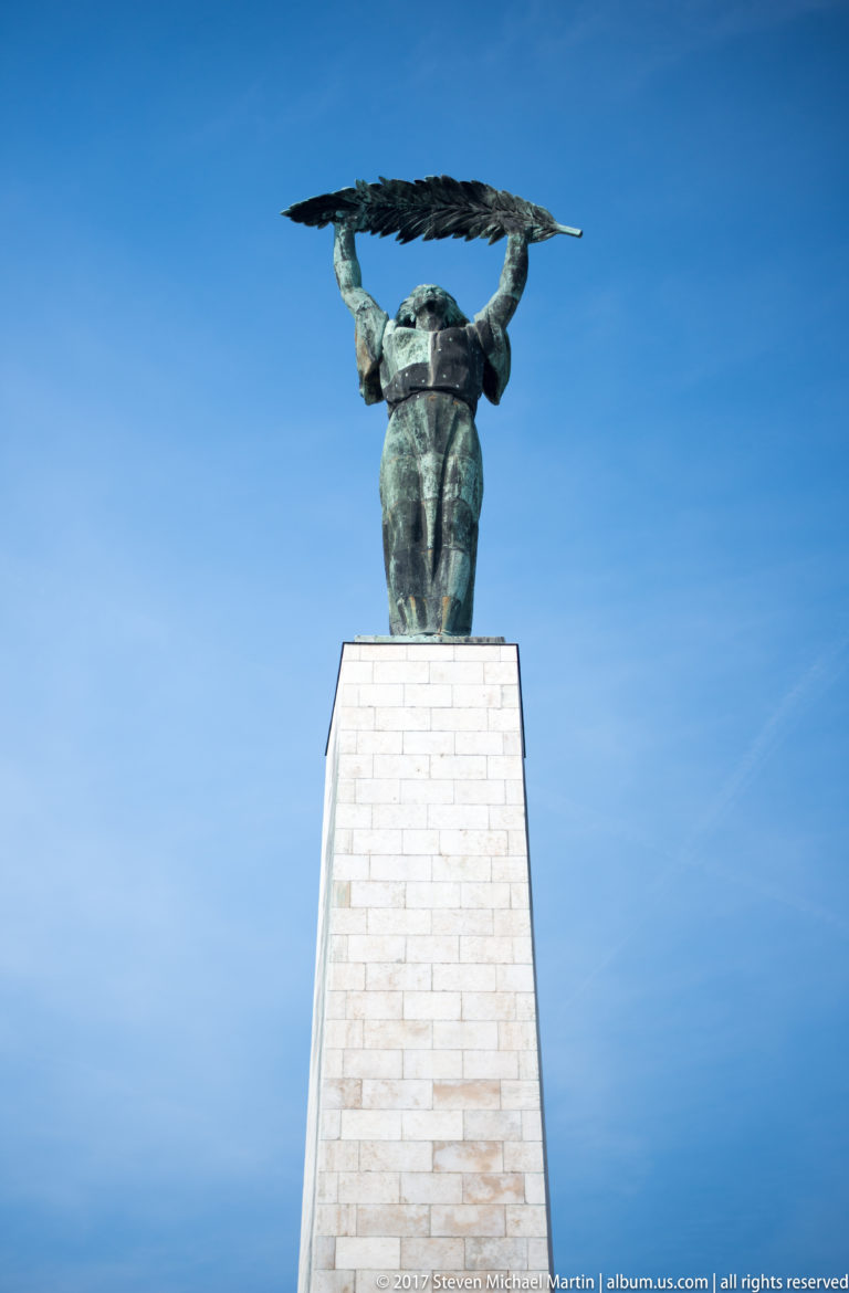 Full low-angle view of Budapest's Liberty Statue and tall pale stone pedestal beneath a blue sky.
