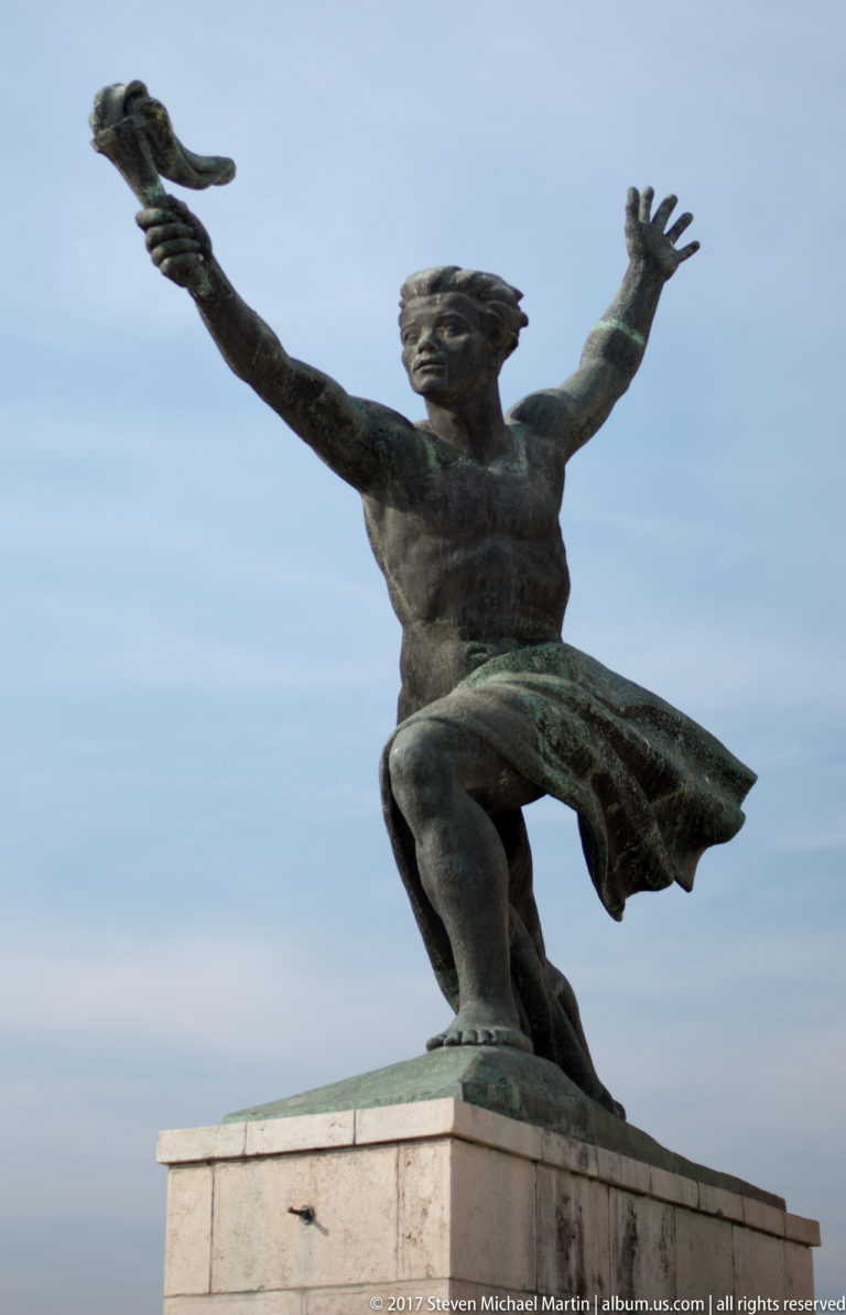 Bronze male figure with arms spread wide and drapery sweeping behind him on a stone plinth.
