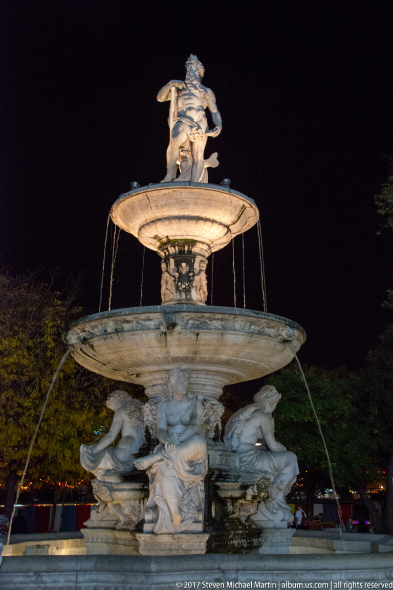 Illuminated multi-tiered stone fountain with reclining figures around the base and a sculptural group at the top.