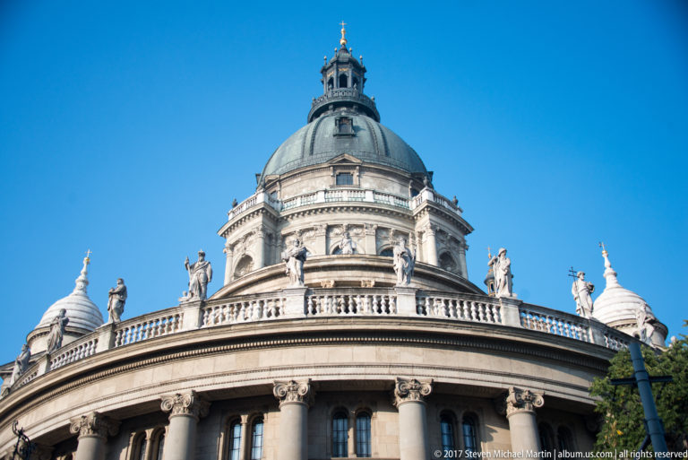Close low-angle view of St. Stephen’s Basilica dome, lantern, curved colonnade, and rooftop statues against blue sky.