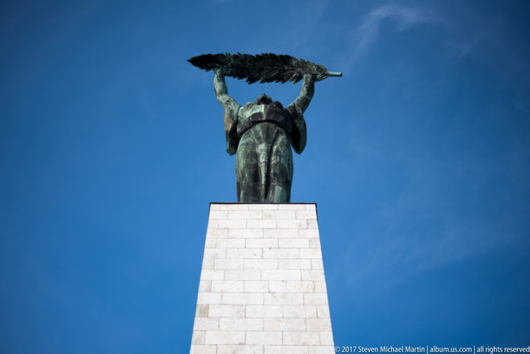 Budapest Liberty Statue on a tall stone pedestal, female figure raising a palm frond overhead against blue sky.