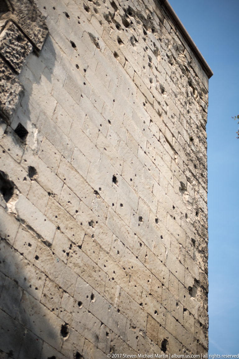 Angled close-up of a pale stone Citadella wall with small openings against a clear blue sky.