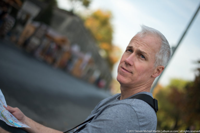 Gray-haired man holding a folded map and looking upward, with a blurred pedestrian area behind him.
