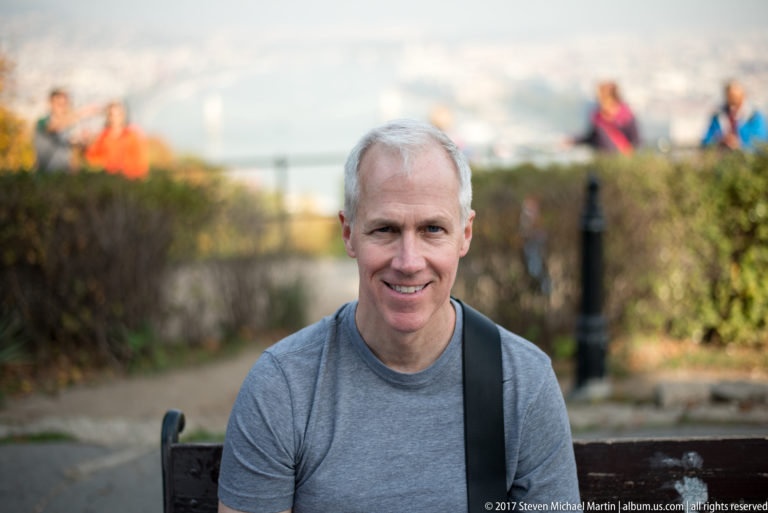 Gray-haired man seated on a bench at a Budapest overlook, with hedges and visitors blurred behind him.
