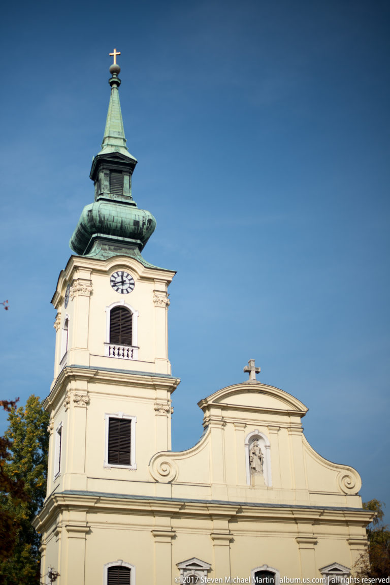 Pale Baroque church with a single clock tower, green roof, clock face, cross, and curved gable.