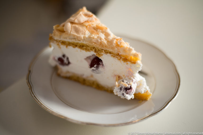Slice of layered cream cake with cherries and a meringue-like topping on a white plate.