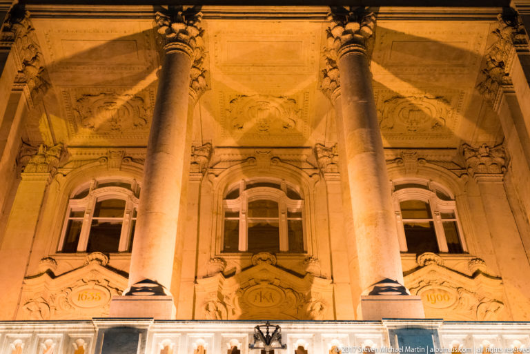 Illuminated stone façade with giant columns, arched windows, sculpted masks, and carved date cartouches.
