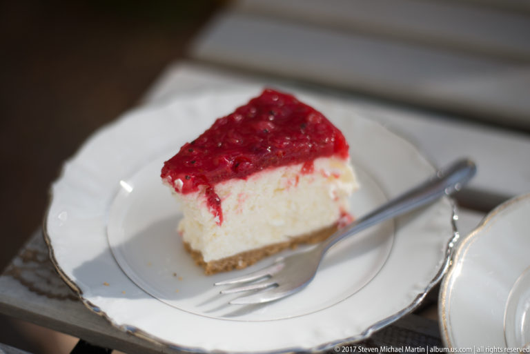 Square slice of cheesecake with glossy red berry topping on a white plate beside a fork and spoon.