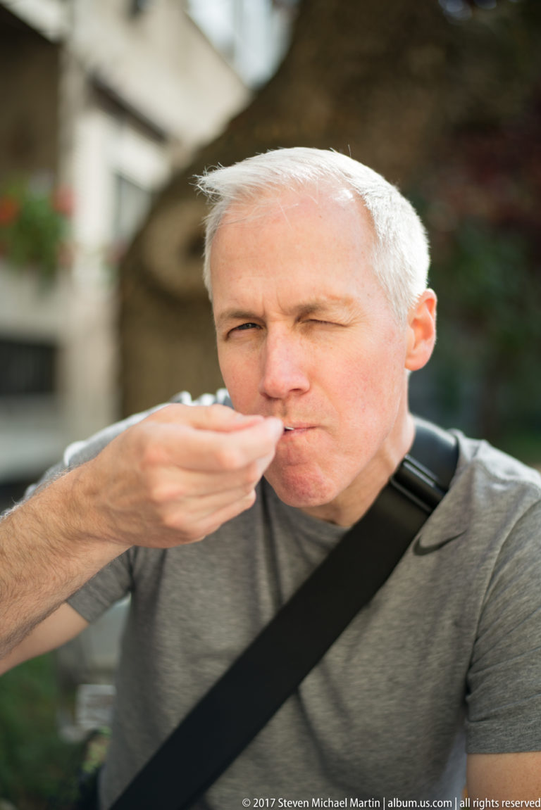 Gray-haired man outdoors taking a bite from a spoon, with the background softly blurred.
