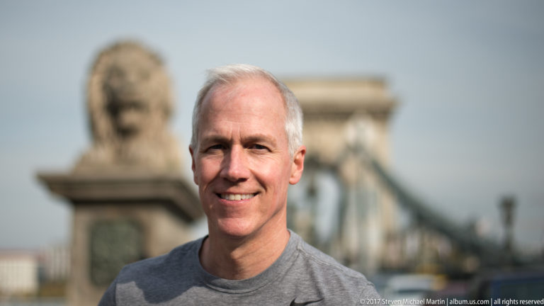 Gray-haired smiling man with a Chain Bridge lion and stone bridge tower blurred in the background.