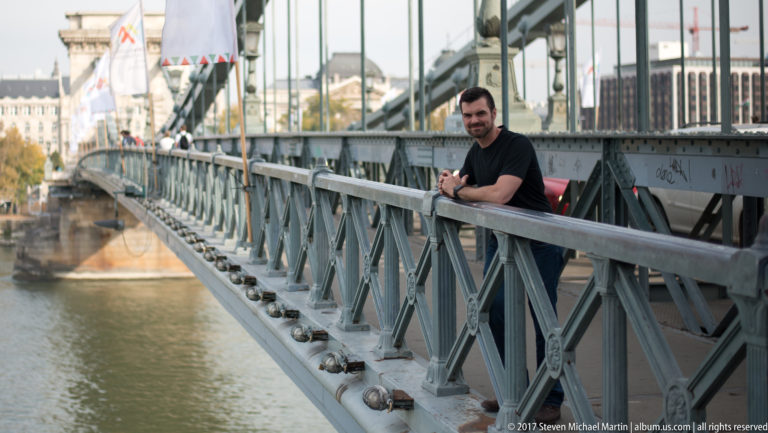 Dark-haired man leaning on a riveted metal bridge railing above the Danube, with bridge structure receding behind.