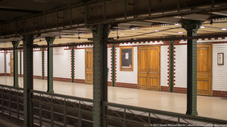 Historic metro platform with white and red-brown tiles, green iron columns, decorative railings, and wooden doors.