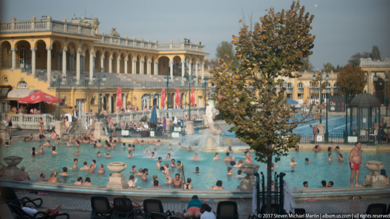 Crowded turquoise outdoor thermal pool at Széchenyi Baths surrounded by yellow arcades and autumn trees.