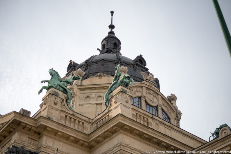 Low-angle view of Széchenyi Baths corner dome with green-patinated rooftop figures and ornate pale stone façade.
