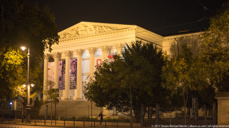 Illuminated Neoclassical museum façade with tall columns, sculpted pediment, broad steps, and exhibition banners.
