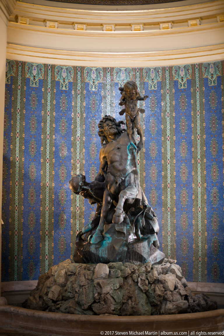 Patinated bronze group of intertwined human figures on a rocky base before a patterned blue-green tiled wall.