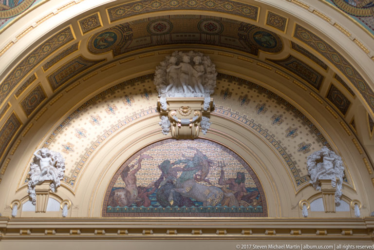 Ornate interior arch with a large figurative mosaic, sculpted figures, gilded details, and patterned ceiling decoration.