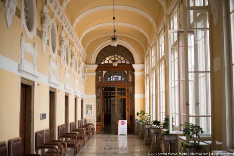 Long yellow-vaulted corridor with tall windows, decorative wall reliefs, brown chairs, and a wooden doorway at the far end.