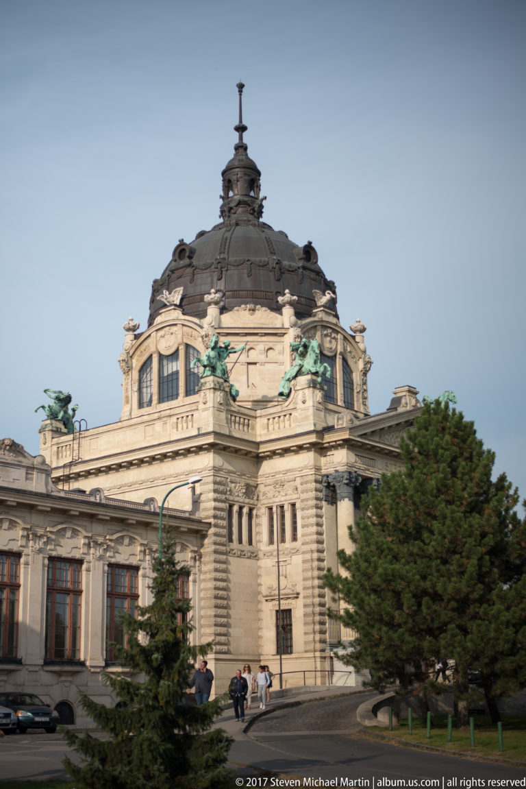 Ornate pale stone building with dark domed roof, sculptural decoration, and green-patinated figures beside trees.