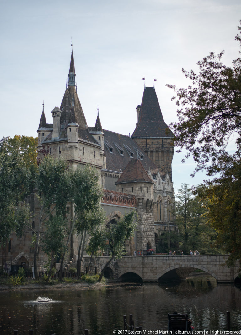 Vajdahunyad Castle across its moat with stone towers, steep roofs, trees, and a small bridge.