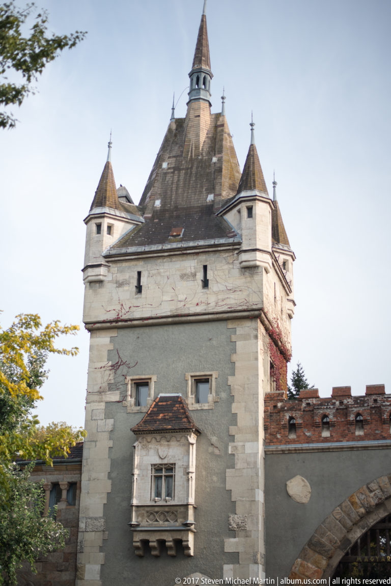 Square stone tower with steep roof, crenellated wall, and arched gateway at Vajdahunyad Castle.