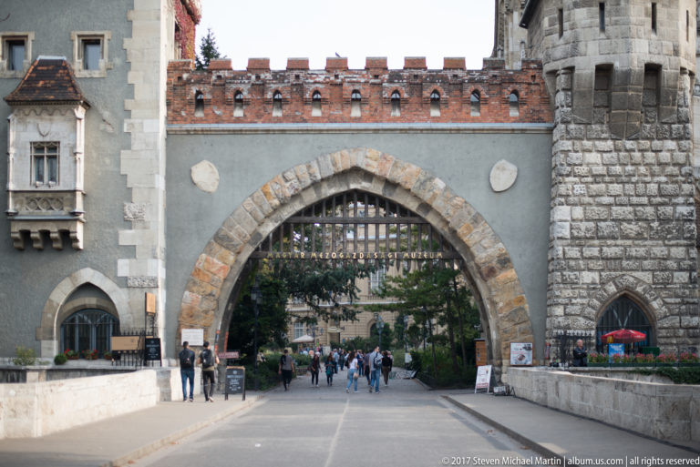 Pointed-arch gateway at Vajdahunyad Castle with crenellated walls, towers, and visitors walking through.