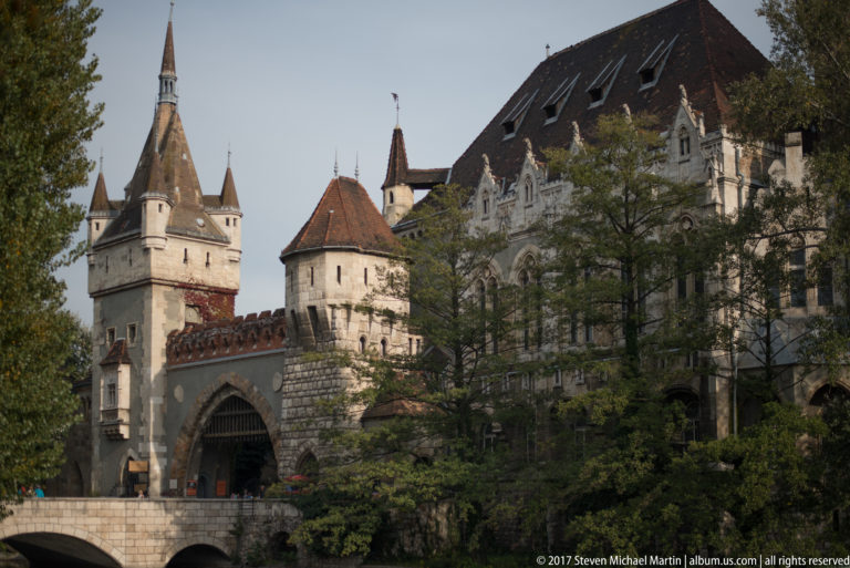 Oblique view of Vajdahunyad Castle showing gateway, conical towers, ornate façades, trees, and the moat.