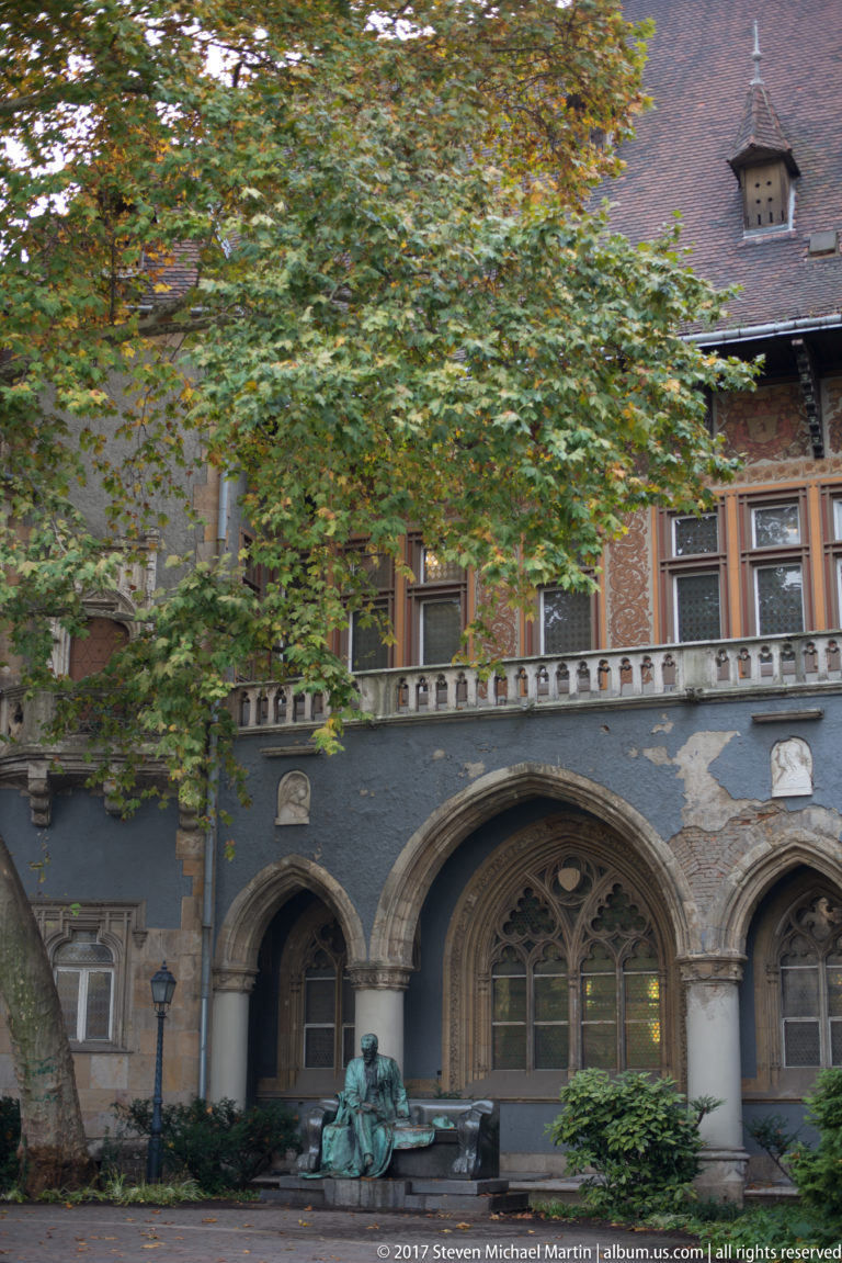 Arcaded stone façade with traceried windows and a seated bronze figure beneath trees at Vajdahunyad Castle.