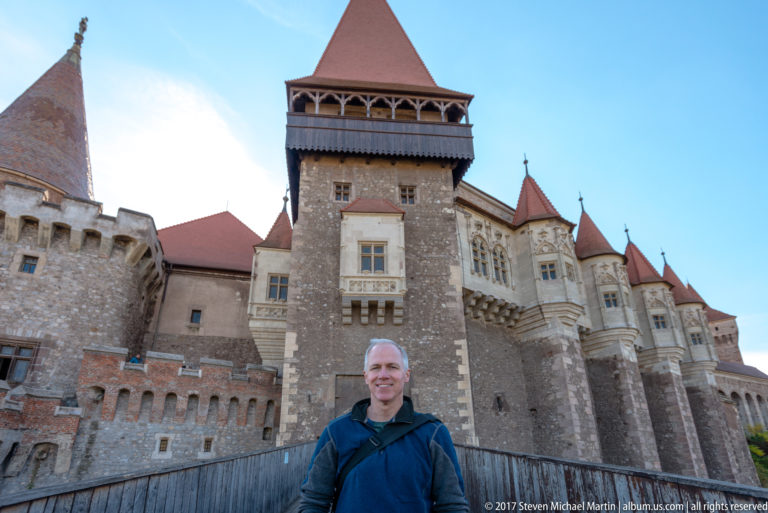 Man standing on Corvin Castle’s entrance bridge with the gate tower and stone façade behind him.