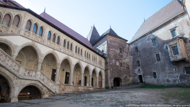 Corvin Castle courtyard with two-level arcade, exterior staircase, pointed windows, and surrounding stone walls.