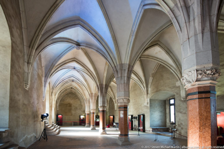 Large Corvin Castle hall with Gothic rib vaults, stone columns, carved capitals, and museum displays.